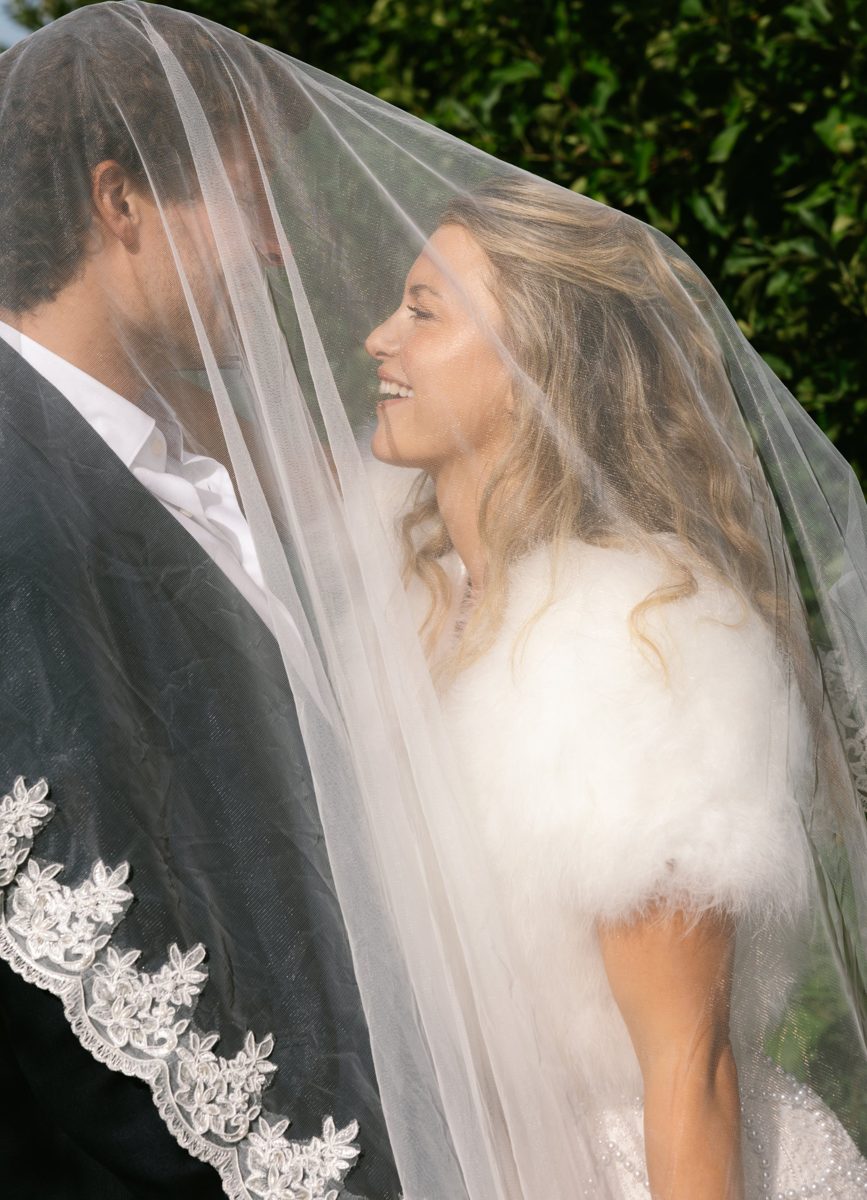 Bride and groom smiling under a veil in front of green hedges