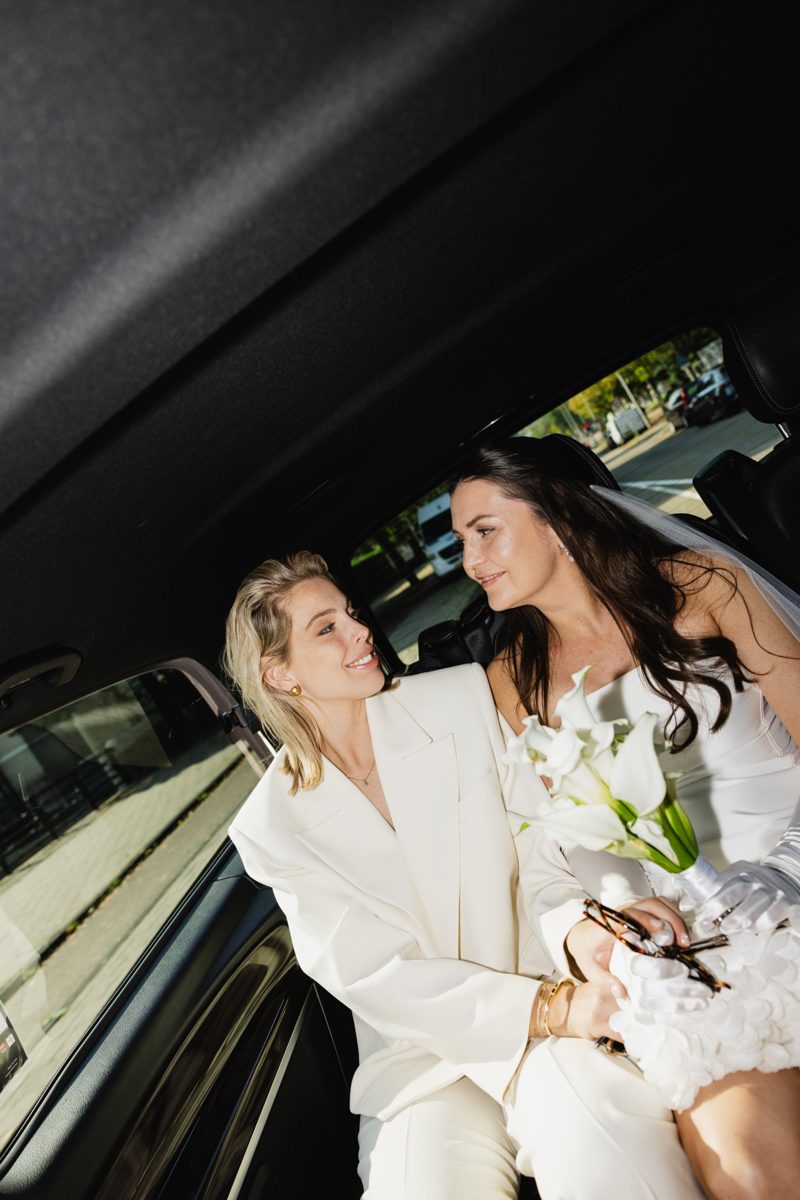 Two brides smiling at each other in the back of a car, lit by flash